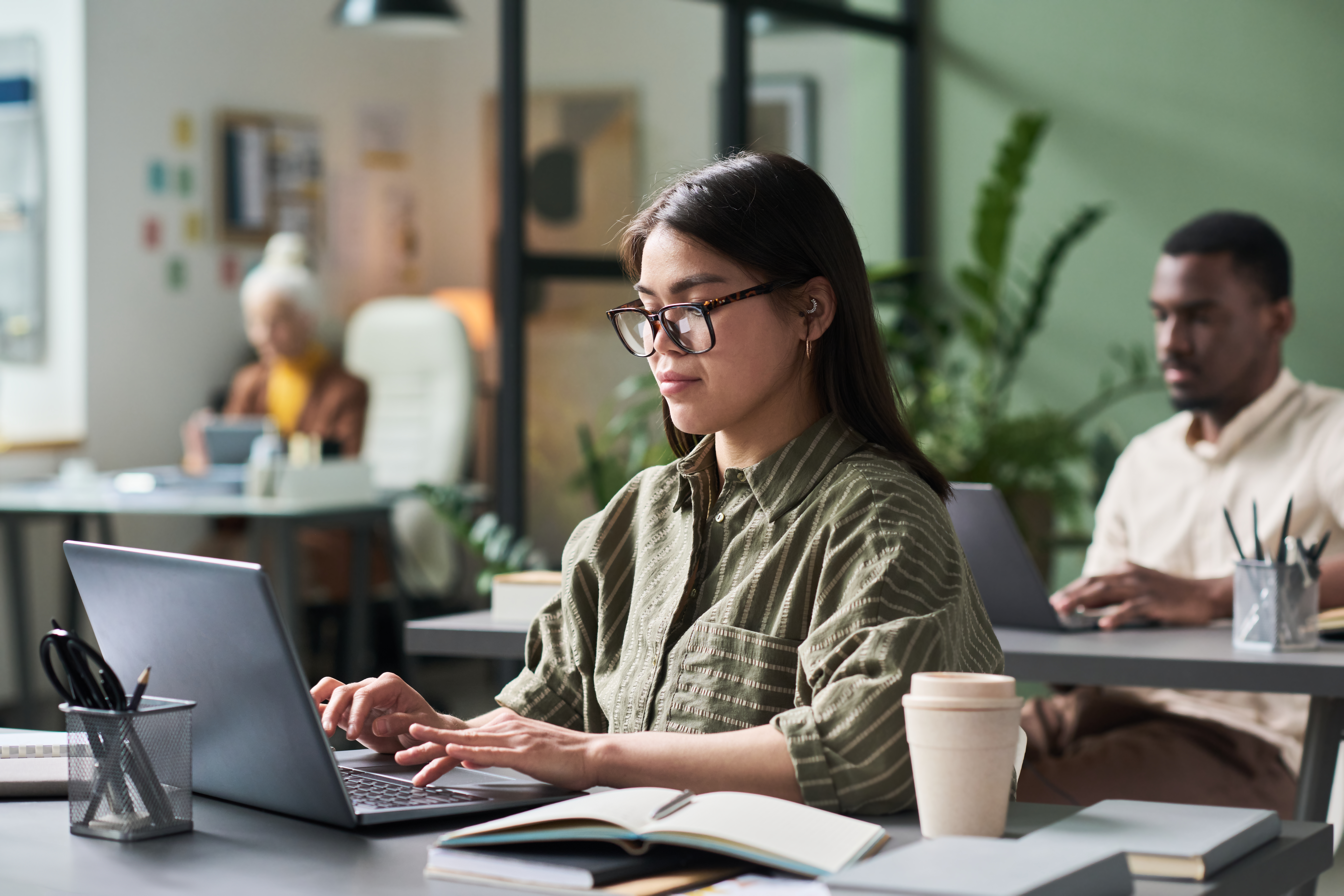 A focused woman types on a laptop in a modern office, surrounded by colleagues also working on computers. The environment reflects a collaborative setting where AI tools are likely being used to streamline tasks such as the financial close process.