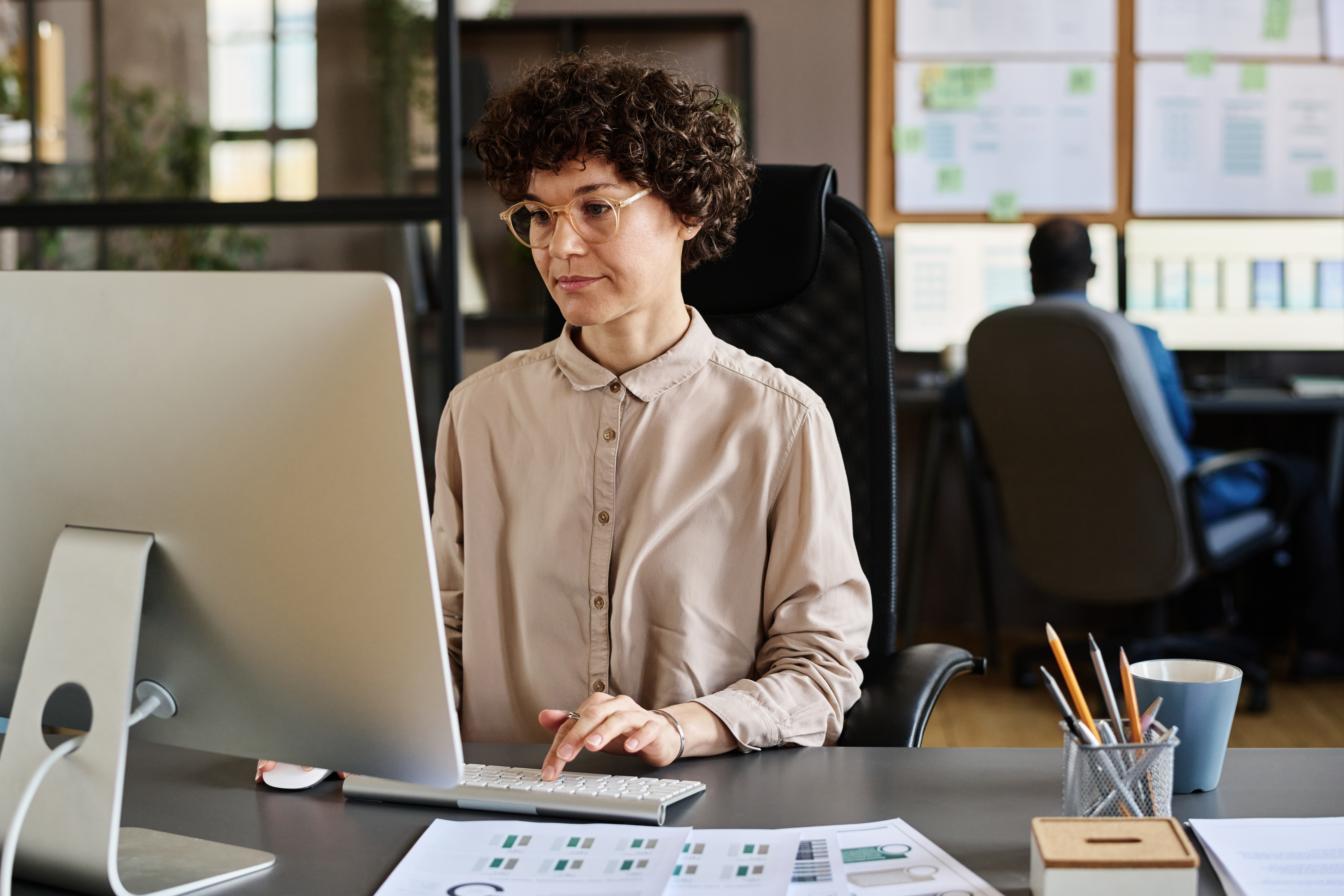 A professional woman works intently on a desktop computer in a modern office, analyzing financial data with charts visible on her desk. Her focused demeanor suggests she is using AI tools to support a streamlined financial close process. The background features additional screens and planning boards, reinforcing a data-driven work environment.
